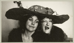 Two women sit under a giant hat, one pulls a funny face, both look mischeivous